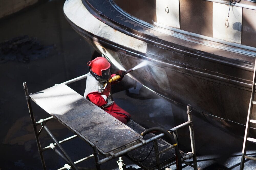 Worker cleaning hull of boat with high pressure hose in shipyard